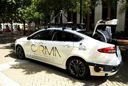 A car with its trunk open is shown. The car has the CARMA logo on its side, along with the USDOT FHWA logo and the characeteristic honeycomb icons associated with CARMA. The car is also equipped with various electronic equipment affixed to its roof and front and rear bumpers.