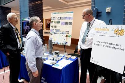 Two men stand in front of a table and one man stands to the side of the table. The table has various pavement materials on it, and a poster is located behind the table.