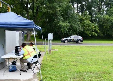 Two workers in yellow vests sit at a table outside and under a tent. Computer equipment is located on the table, and a sport utility vehicle travels on the road in front of the workers.