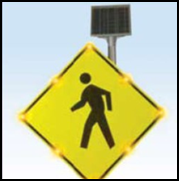 The photograph shows an icon of a walking pedestrian in a diamond-shaped highway sign. The sign has seven lights shown that are embedded onto the edge of the sign. A solar panel is located above the sign.
