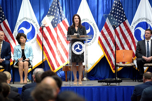 A woman stands at a podium and gives a speech to attendees who are not seen in the photo. A man and a woman are seated to the woman's left, and a man is seated to her right.