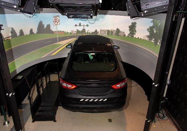 This photo shows a black car with a virtual reality screen in front of it. The screen shows an image of a road with a 'no u-turn' sign and trees on either side.
