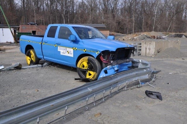 This photo shows a blue pickup truck with its frontend smashed. There is a metal road barrier in front of the pickup truck indicating the pickup truck ran into the barrier. The barrier is bent, but still in place.