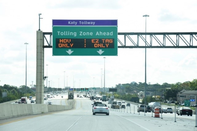 A sign on a highway says 'Katy Tollway' and 'Tolling Zone Ahead.' Underneath on the same sign, electronic text says HOV Only on one side and EZ TAG Only. The freeway shows cars going both directions with a concrete barrier between them.
