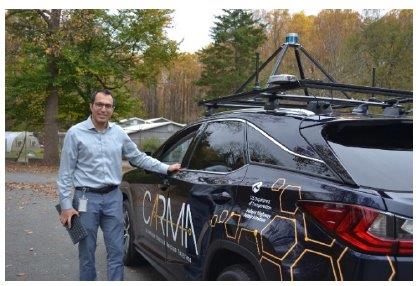 A man looks at the camera while standing next to an SUV. The vehicle has equipment affixed to its roof.