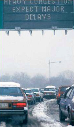 Photo: Variable message sign reporting traffic delays wth cars driving below on icy and snowy roads.