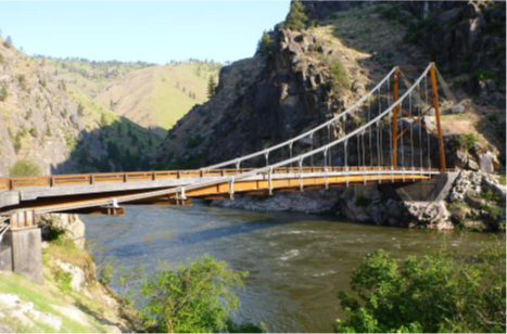 This is a photo of a one-span bridge over a river in the mountains of Oregon. No vehicles or people are on the bridge. The sky is clear. It is sunny.
