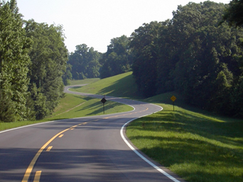 This is a photo of a winding section of the Natchez Trace Parkway. There are no cars on the road. It's a sunny day with dense forests on each side of the road beyond grassy shoulders.