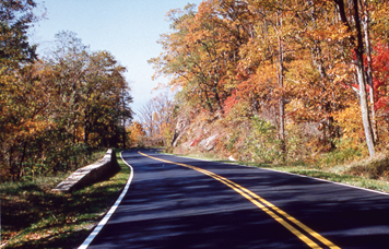 This is a photograph of a road in Shenandoah National Park It is Fall. The leaves on each side of the road are orange, brown, yellow, and red. No cars are on the road.