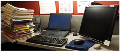 Photo of a desk with a high pile of paperwork, a laptio, mouse, and monitor.