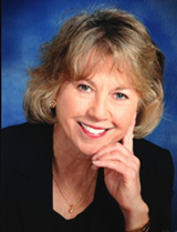 Title: Kathy Facer - Description: Headshot of women in black suit in front of blue background.
