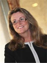 Title: Lisa Harrison - Description: Headshot of woman black suit in front of yellow background.