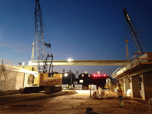 Picture of the construction of a highway overpass. Two cranes are lifting a concrete slab into position. The photo was taken around dusk.