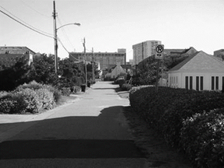 Un vecindario - Description: Esta imagen en blanco y negro es de una acera en un barrio residencial. Un letrero de 'No Estacione' se encuentra a la izquierda de la acera al lado de una casa.