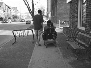 Dos personas en una acera - Description: Esta es una foto en blanco y negro de un peatón y una persona con movilidad reducida en una acera al lado de un portabicicletas. Sus espaldas están hacia la cámara.