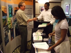 Photo of Paul Tufts, FHWA Environmental Program Specialist standing in front of the Resource Center exhibit booth while he, shakes hands with a conference attendee. A second person stands nearby, looking over materials on the exhibit table.