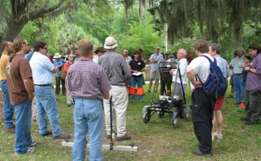 Photo of Dr. Lew Somers leading a discussion of geophysical techniques used during field investigations. Workshop participants are standing around him watching as he speaks.