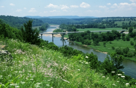 Photo of US Scenic Highway 62, Marion Baxter County, AR. The photo features a bridge portion of the highway crossing over a river that is surrounded by rolling hills covered with trees, grasses and wild flowers.
