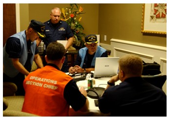 Photo of a group of incident responders sitting around an office table spread with papers. One individual is wearing an orange vest that reads 'Operations Section Chief.'