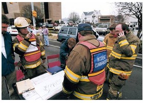 Photo of a group of responders in an urban area surrounding a table with documents spread out on it. Two responders near another wearing a vest labeled 'Incident Commander' are communicating over walkie talkie.