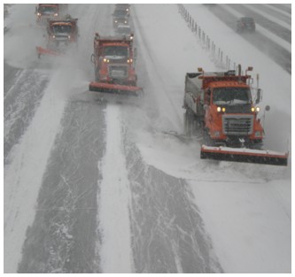 Photograph of a line of snowplows, each plowing a lane on a multi-lane interstate highway.